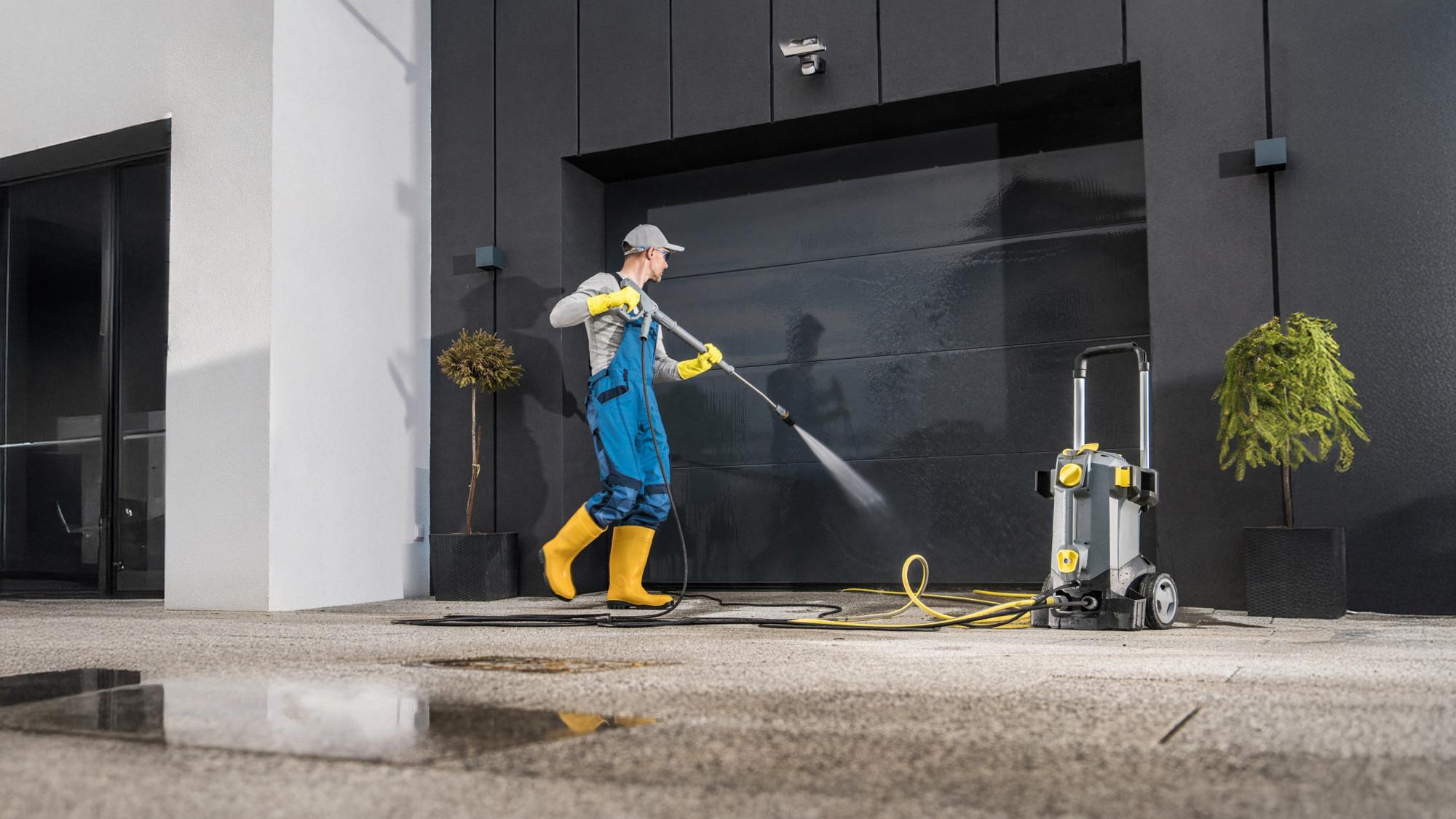 A person in a gray cap, blue overalls, and yellow rubber boots is using a pressure washer on a driveway near a modern garage. The pressure washer is on the ground beside them, with a hose connected. There are potted plants nearby and wet spots on the pavement.